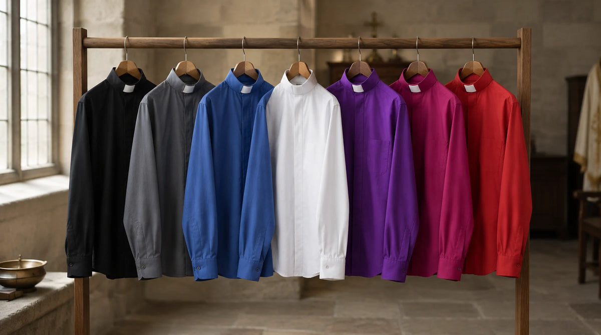Seven clergy shirts with white tab collars hanging on a wooden rail in a stone sacristy: black, grey, blue, white, violet purple, amaranth and scarlet