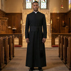 A minister wearing the black cassock robe with its sash belt, standing in the centre aisle of a stone church.