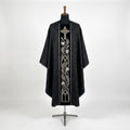 Front of a black damask chasuble with a black velvet column orphrey embroidered in silver with a cross, a lily vine and gothic arches