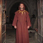 A man wearing a burgundy monk habit with the hood raised, the gold sunburst embroidery visible on the chest.