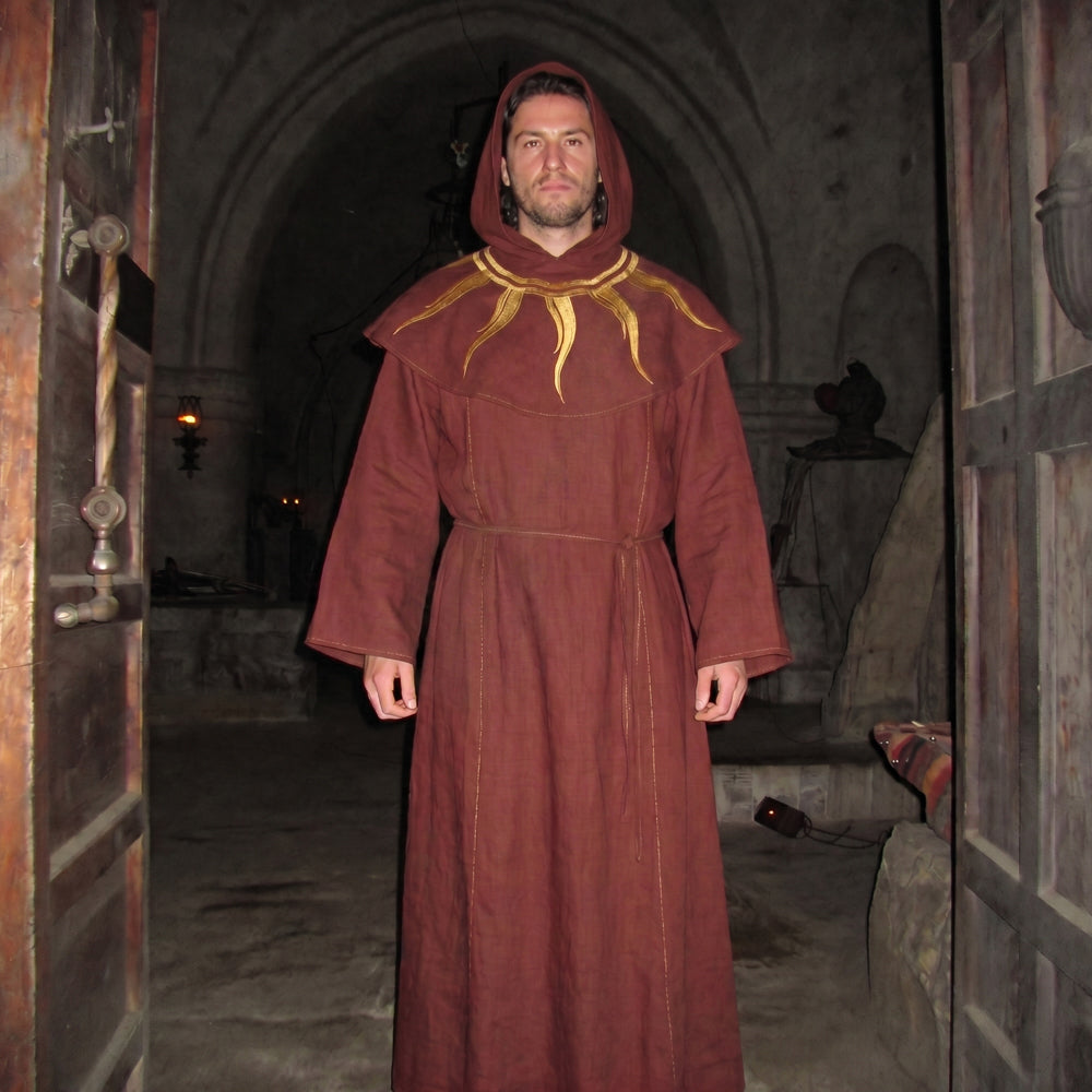 A man wearing a burgundy monk habit with the hood raised, the gold sunburst embroidery visible on the chest.