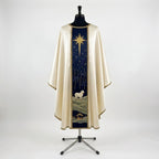 Front of an ivory satin Nativity chasuble with a navy column orphrey: a gold star with long rays above a white lamb on green hills and the stable at the hem